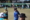 Woman lawn bowls player in a blue uniform and visor raises her arm in celebration on a sunny outdoor green, with other bowlers and bowls visible in the background